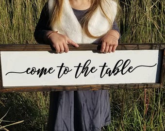 May include: A white wooden sign with black lettering that reads "come to the table". The sign is held by a person in front of a grassy background.