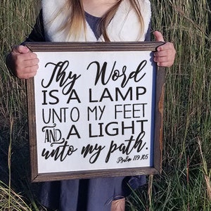 May include: A framed wooden sign with the text "Thy Word is a Lamp Unto My Feet And a Light Unto My Path." The sign has a white background with black lettering and a dark brown frame. The sign is held in front of a field of tall grass.