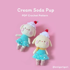 Op de afbeelding: Twee gehaakte amigurumi-speelgoedjes in de vorm van een room-soda-puppy. De speelgoedjes dragen blauwe en witte jurken met witte stippen. Een speelgoedje heeft een gele sjaal. De speelgoedjes hebben rode kersaccenten op hun hoofd. De afbeelding bevat de tekst "Cream Soda Pup" en "PDF Crochet Pattern".