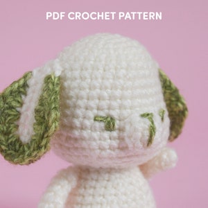 Op de afbeelding: Een wit gehaakt amigurumi-speelgoed met groene oren. Het speelgoed heeft een eenvoudig gezicht met groene ogen en een kleine neus.
