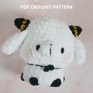 May include: White crocheted plush toy with black and yellow horns, black eyes, and a black bow tie. The toy has black spots on its legs and is sitting on a pink surface.