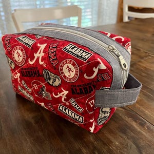Könnte beinhalten: Eine rot-weiß gesteppte Kosmetiktasche mit dem Logo und Text der University of Alabama Crimson Tide. Die Tasche hat einen grauen Reißverschluss und einen passenden Griff. Die Tasche ist rechteckig und liegt auf einer Holzoberfläche.