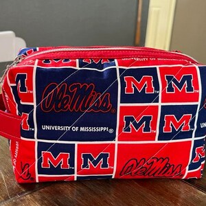 Peut inclure: Une trousse de maquillage matelass&eacute;e rouge, blanche et bleue avec le logo Ole Miss et le nom de l'Universit&eacute; du Mississippi. Le sac a une fermeture &eacute;clair rouge et des poign&eacute;es rouges. Le sac est de forme rectangulaire.