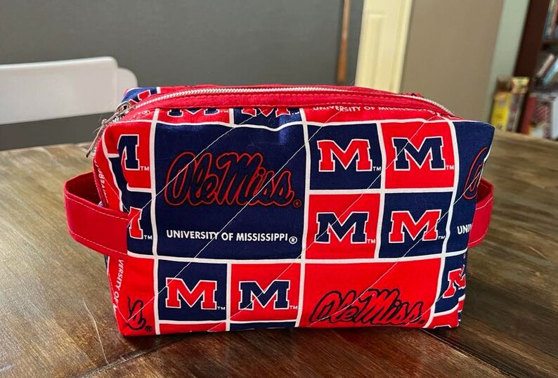 Peut inclure: Une trousse de maquillage matelass&eacute;e rouge, blanche et bleue avec le logo Ole Miss et le nom de l'Universit&eacute; du Mississippi. Le sac a une fermeture &eacute;clair rouge et des poign&eacute;es rouges. Le sac est de forme rectangulaire.