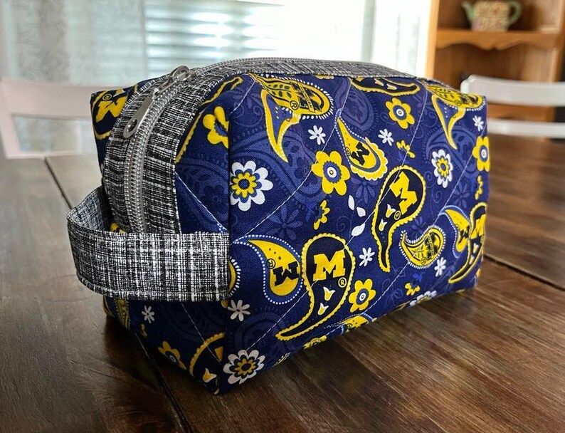Peut inclure: Une trousse de toilette rectangulaire matelass&eacute;e avec un motif cachemire bleu marine, des accents floraux jaunes et la lettre "M". Le sac a une fermeture &eacute;clair et une poign&eacute;e &agrave; carreaux noirs et blancs. Environ 20 cm de long, 13 cm de large et 10 cm de haut.