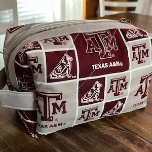 Peut inclure: Une trousse de toilette rectangulaire grise et bordeaux, avec une fermeture &eacute;clair et une petite poign&eacute;e. Le sac pr&eacute;sente un motif r&eacute;p&eacute;t&eacute; du logo Texas A&M et les mots "TEXAS A&M". Le sac est en tissu matelass&eacute;.
