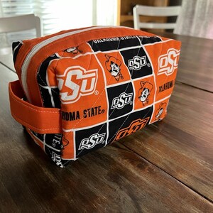 Peut inclure: Trousse de maquillage matelass&eacute;e orange et noire avec le logo et le texte de l'Oklahoma State University. Le sac a une fermeture &eacute;clair, une poign&eacute;e orange et une forme rectangulaire. Le logo OSU est r&eacute;p&eacute;t&eacute;.