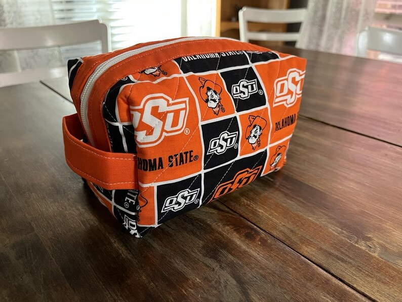 Peut inclure: Trousse de maquillage matelass&eacute;e orange et noire avec le logo et le texte de l'Oklahoma State University. Le sac a une fermeture &eacute;clair, une poign&eacute;e orange et une forme rectangulaire. Le logo OSU est r&eacute;p&eacute;t&eacute;.