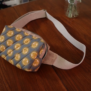 Puede incluir: Una riñonera acolchada gris y amarilla con una correa ajustable beige. La riñonera tiene un patrón repetitivo de soles amarillos con caras rosas.