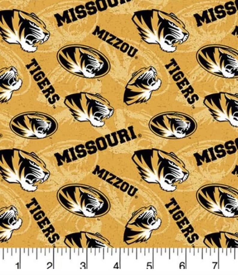 Peut inclure: Tissu dor&eacute; avec un motif r&eacute;p&eacute;t&eacute; de t&ecirc;tes de tigre noires et dor&eacute;es, les mots "Tigers", "Missouri" et "Mizzou". Le tissu a une r&egrave;gle en bas avec des mesures en pouces.
