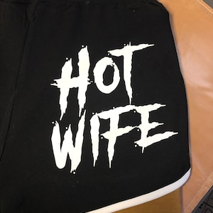 May include: Black athletic shorts with a white graphic that says "HOT WIFE" in a stylized font.