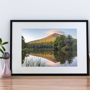 Può includere: Fotografia incorniciata di un paesaggio con una montagna riflessa in un lago. L'immagine è in una cornice nera e poggia su una superficie di legno. Un vaso bianco con vegetazione, una sfera decorativa e libri sono anch'essi presenti.