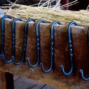 May include: Seven black metal hooks with a twisted design are mounted on a wooden surface. The hooks are all the same size and shape, and they are arranged in a row.