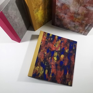 Könnte beinhalten: Eine Sammlung von Kunstjournalen mit farbenfrohen abstrakten Designs. Die Journale haben goldfarbene Rücken und Einbände mit roten, blauen und gelben Farbspritzern. Andere Journale haben graue, goldene und kastanienbraune Einbände.