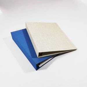 Könnte beinhalten: Zwei Ordner. Einer ist einfarbig blau, der andere hat eine neutrale beige Farbe. Die Ordner sind rechteckig mit einem Drei-Ring-Mechanismus. Die Ordner bestehen aus einem strukturierten Material.