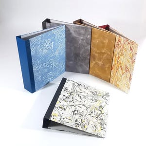 Può includere: Una collezione di cinque raccoglitori ad anelli con vari design di copertina. Quattro raccoglitori sono disposti in piedi in fila, mostrando motivi marmorizzati blu, grigi, dorati e gialli. Un raccoglitore è appoggiato in piano in primo piano con una copertina marmorizzata nera, bianca e gialla.