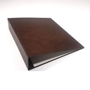 Può includere: Una scatola rettangolare marrone scuro con un piano leggermente inclinato. La scatola ha un design minimalista e sembra essere fatta di legno o materiale simile. L'interno è visibile, rivelando carta bianca.