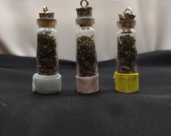 May include: Three small glass bottles with cork tops and silver caps. Each bottle is filled with dried herbs and has a different colored fabric covering the bottom. The bottles are arranged in a row on a black surface.