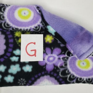 May include: Purple and black fleece blanket with a floral pattern. The blanket is folded in half and has a purple fleece lining. The letter "G" is written on a white piece of paper.