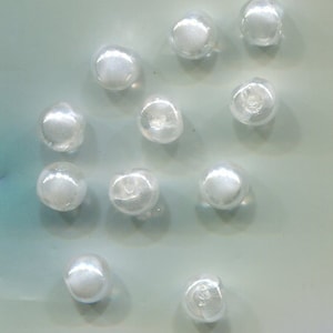 Peut inclure: Douze perles rondes en verre blanc, certaines avec un léger reflet irisé, disposées sur une surface bleu clair.