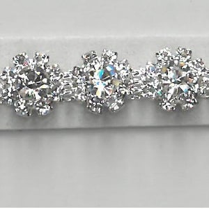 May include: A sparkling silver bracelet featuring a repeating pattern of clear, round-cut gemstones. Each gemstone is surrounded by smaller stones, creating a floral or snowflake-like design. The bracelet is set against a neutral background.