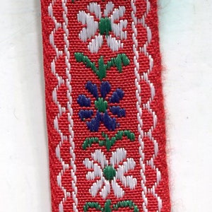 Könnte beinhalten: Roter Stoffbesatz mit einem weißen, grünen und blauen Blumenmuster. Der Besatz hat einen gewellten Rand.