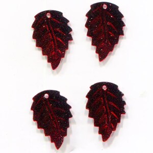 May include: Four red glitter leaf-shaped charms with a small hole at the top for stringing. The leaves have a dark red outline and a lighter red interior with vein details. The charms are on a white background.