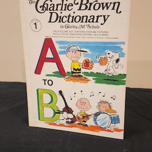 May include: The Charlie Brown Dictionary, Volume 1, features a red letter A and illustrations of Charlie Brown, Snoopy, and other Peanuts characters. The book cover includes the text "The 8 volume set contains over 600 pictures in full color. 2500 words defined. 50 U.S. maps."