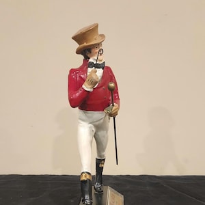 K&ouml;nnte beinhalten: Figur eines Herrn in roter Jacke, wei&szlig;er Hose und schwarzen Stiefeln, der einen Gehstock und ein Monokel h&auml;lt. Er tr&auml;gt einen braunen Zylinder und eine Fliege. Auf dem Sockel der Figur steht der Text "JOHNNIE WALKER".
