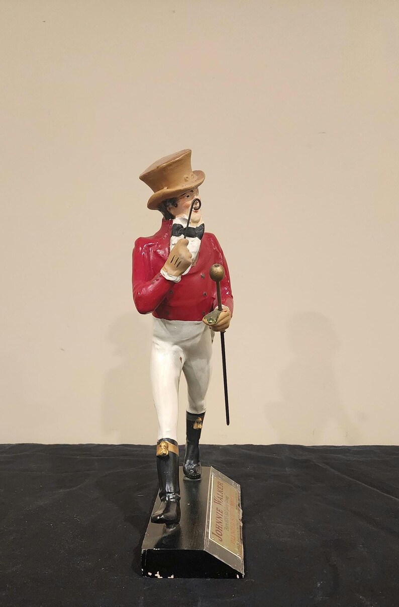 K&ouml;nnte beinhalten: Figur eines Herrn in roter Jacke, wei&szlig;er Hose und schwarzen Stiefeln, der einen Gehstock und ein Monokel h&auml;lt. Er tr&auml;gt einen braunen Zylinder und eine Fliege. Auf dem Sockel der Figur steht der Text "JOHNNIE WALKER".