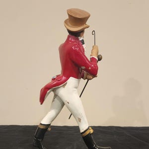 K&ouml;nnte beinhalten: Dekorative Figur eines Mannes in rotem Mantel, wei&szlig;er Hose und schwarzen Stiefeln, der einen Stock h&auml;lt. Die Figur tr&auml;gt einen braunen Zylinder und ist in einer Laufhaltung dargestellt. Die Figur ist auf einem schwarzen rechteckigen Sockel montiert.