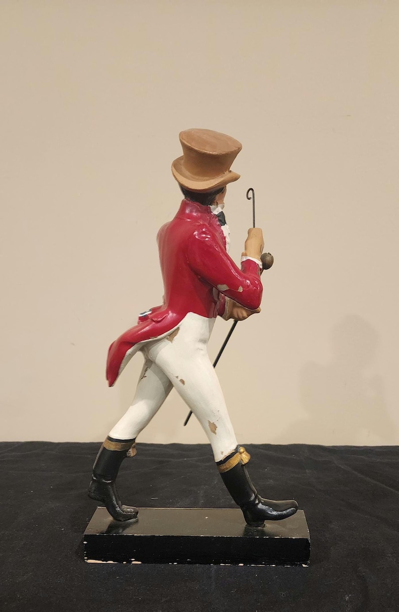 K&ouml;nnte beinhalten: Dekorative Figur eines Mannes in rotem Mantel, wei&szlig;er Hose und schwarzen Stiefeln, der einen Stock h&auml;lt. Die Figur tr&auml;gt einen braunen Zylinder und ist in einer Laufhaltung dargestellt. Die Figur ist auf einem schwarzen rechteckigen Sockel montiert.