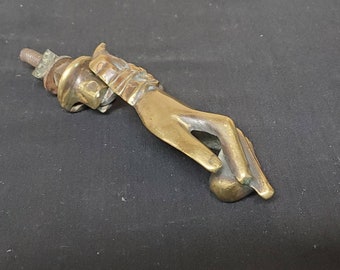 May include: A brass door knocker in the shape of a hand holding a sphere. The knocker is mounted on a wall.
