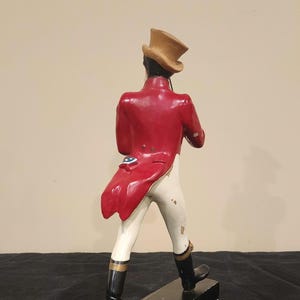 K&ouml;nnte beinhalten: Vintage Figur einer Person in rotem Mantel, wei&szlig;er Hose und schwarzen Stiefeln, die einen beigen Zylinder tr&auml;gt. Die Figur steht auf einem schwarzen Sockel. Der Mantel hat ein dekoratives blau-wei&szlig;es Emblem.