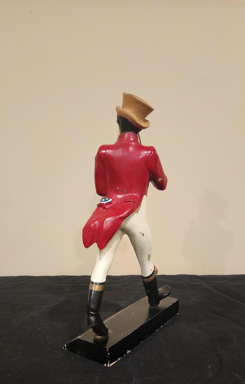 K&ouml;nnte beinhalten: Vintage Figur einer Person in rotem Mantel, wei&szlig;er Hose und schwarzen Stiefeln, die einen beigen Zylinder tr&auml;gt. Die Figur steht auf einem schwarzen Sockel. Der Mantel hat ein dekoratives blau-wei&szlig;es Emblem.