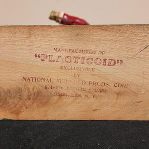 Puede incluir: Un bloque de madera con el texto "MANUFACTURED OF 'PLASTICOID' EXCLUSIVELY BY NATIONAL MOLDED PRODS. CORP" impreso en rojo. El bloque tiene vetas de madera natural y un peque&ntilde;o objeto con tapa roja.