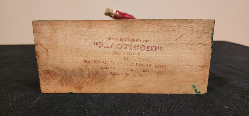 Puede incluir: Un bloque de madera con el texto "MANUFACTURED OF 'PLASTICOID' EXCLUSIVELY BY NATIONAL MOLDED PRODS. CORP" impreso en rojo. El bloque tiene vetas de madera natural y un peque&ntilde;o objeto con tapa roja.