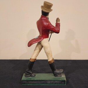 Puede incluir: Figura decorativa de un hombre con abrigo rojo, pantalones beiges y botas negras, sosteniendo un bast&oacute;n. Lleva un sombrero de copa y est&aacute; sobre una base verde. Un objeto de colecci&oacute;n vintage.
