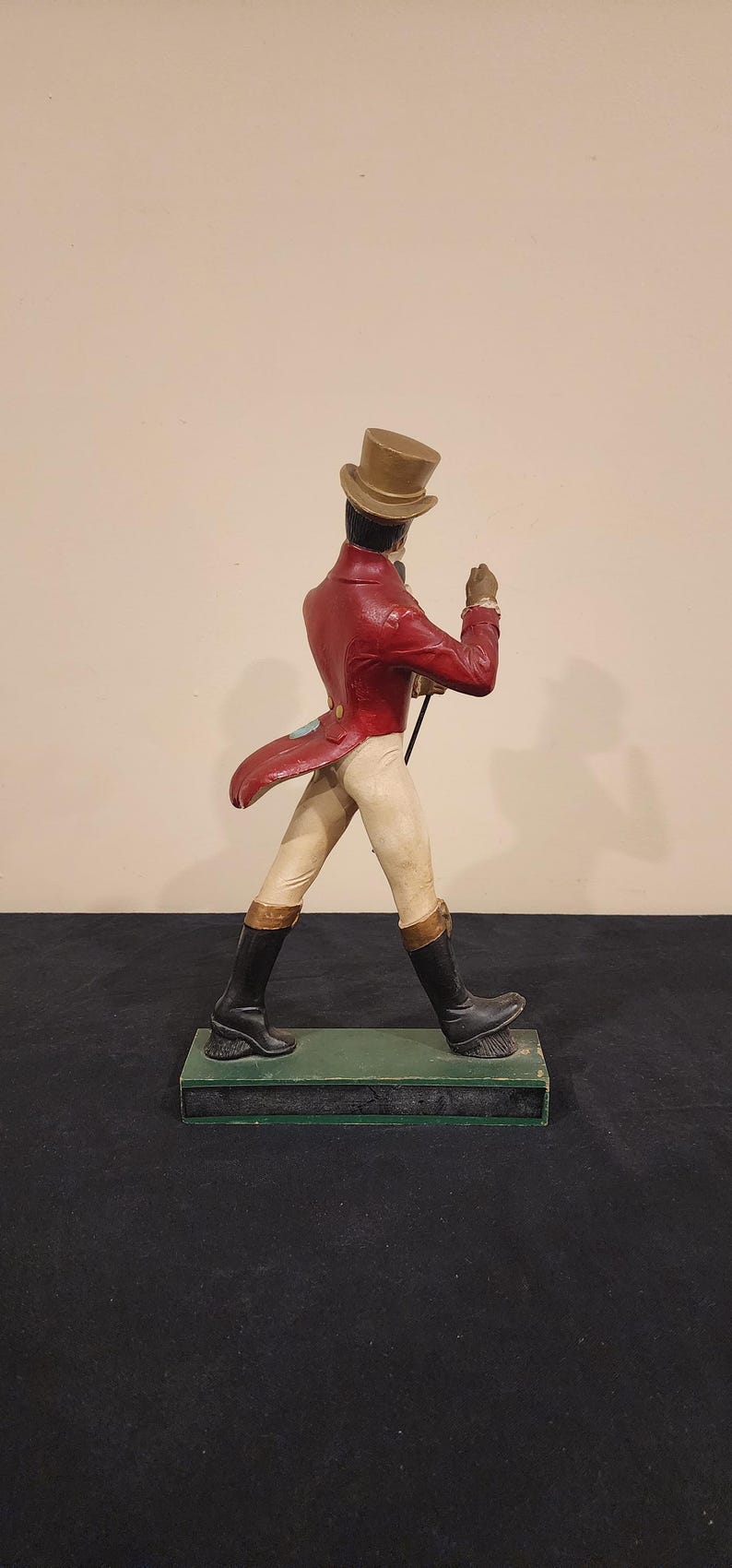 Puede incluir: Figura decorativa de un hombre con abrigo rojo, pantalones beiges y botas negras, sosteniendo un bast&oacute;n. Lleva un sombrero de copa y est&aacute; sobre una base verde. Un objeto de colecci&oacute;n vintage.