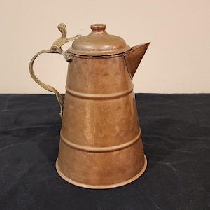 May include: A copper pitcher with a brass handle and lid. The pitcher has a hammered finish and a slightly conical shape.