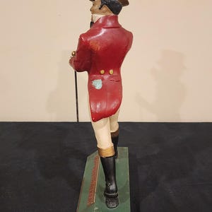Puede incluir: Figura vintage de un caballero con abrigo rojo, botas negras y sombrero de copa, sosteniendo un bast&oacute;n. La figura se alza sobre una base verde con texto. Una pieza decorativa cl&aacute;sica.