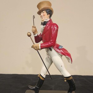 K&ouml;nnte beinhalten: Figur des Johnnie Walker Striding Man, gekleidet in einem roten Mantel, wei&szlig;en Hosen und schwarzen Stiefeln. Die Figur h&auml;lt einen Stock und eine Reitpeitsche. Der Sockel zeigt das Johnnie Walker Logo und Text.