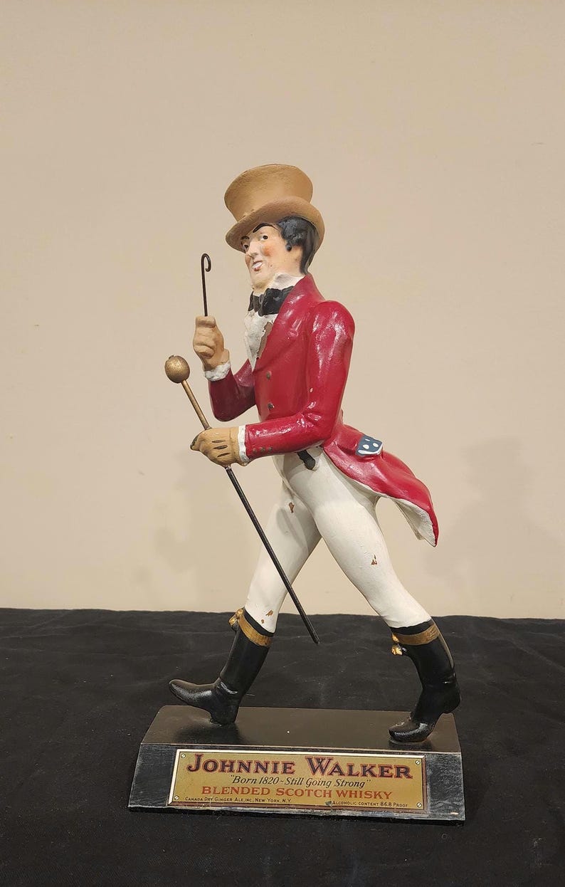 K&ouml;nnte beinhalten: Figur des Johnnie Walker Striding Man, gekleidet in einem roten Mantel, wei&szlig;en Hosen und schwarzen Stiefeln. Die Figur h&auml;lt einen Stock und eine Reitpeitsche. Der Sockel zeigt das Johnnie Walker Logo und Text.