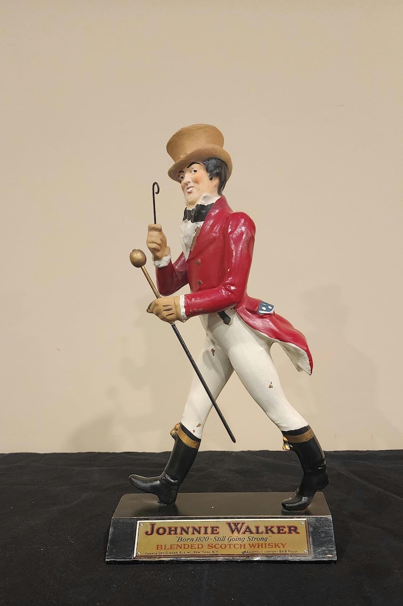 K&ouml;nnte beinhalten: Eine Figur des Johnnie Walker Striding Man, eine Figur in einem roten Mantel, wei&szlig;en Hosen und schwarzen Stiefeln, die einen Stock und ein Monokel h&auml;lt. Auf dem Sockel steht "Johnnie Walker Blended Scotch Whisky".