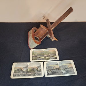 May include: A vintage wooden stereoscope viewer with three antique stereoscopic cards depicting scenes of a mining operation. The viewer has a wooden handle and a metal frame. The cards are printed with images of a mine, a train, and a group of people.