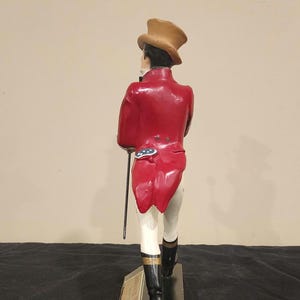 K&ouml;nnte beinhalten: Figur einer Person in rotem Mantel, Zylinder und schwarzen Stiefeln, die einen Stock h&auml;lt. Die Figur steht auf einem schwarzen Sockel mit goldfarbener Aufschrift "JOHNNIE WALKER".