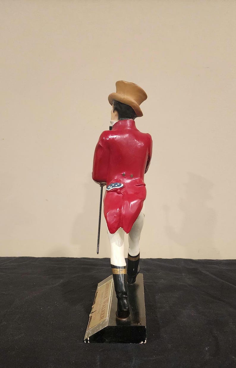K&ouml;nnte beinhalten: Figur einer Person in rotem Mantel, Zylinder und schwarzen Stiefeln, die einen Stock h&auml;lt. Die Figur steht auf einem schwarzen Sockel mit goldfarbener Aufschrift "JOHNNIE WALKER".