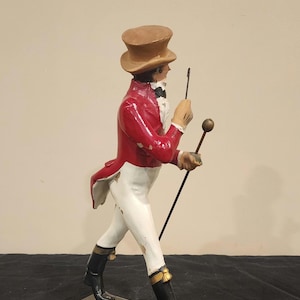 K&ouml;nnte beinhalten: Vintage Figur einer Person in rotem Mantel, wei&szlig;er Hose und schwarzen Stiefeln, die einen Stock und eine Reitpeitsche h&auml;lt. Die Figur tr&auml;gt einen braunen Zylinder und ist auf einem schwarzen rechteckigen Sockel montiert. Dekorationsartikel.