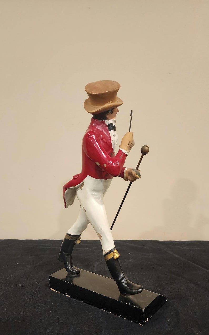 K&ouml;nnte beinhalten: Vintage Figur einer Person in rotem Mantel, wei&szlig;er Hose und schwarzen Stiefeln, die einen Stock und eine Reitpeitsche h&auml;lt. Die Figur tr&auml;gt einen braunen Zylinder und ist auf einem schwarzen rechteckigen Sockel montiert. Dekorationsartikel.