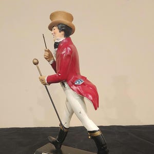 K&ouml;nnte beinhalten: Figur des Johnnie Walker Striding Man. Die Figur tr&auml;gt einen roten Mantel, eine wei&szlig;e Hose und schwarze Stiefel und h&auml;lt einen Stock und einen kleinen Stab. Auf dem Sockel stehen der Markenname und Text.